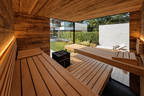 View from the Thermasol Ombra sauna benches through the glass corner onto a garden, with rustic wood walls, light slatted benches and a black stone heater
