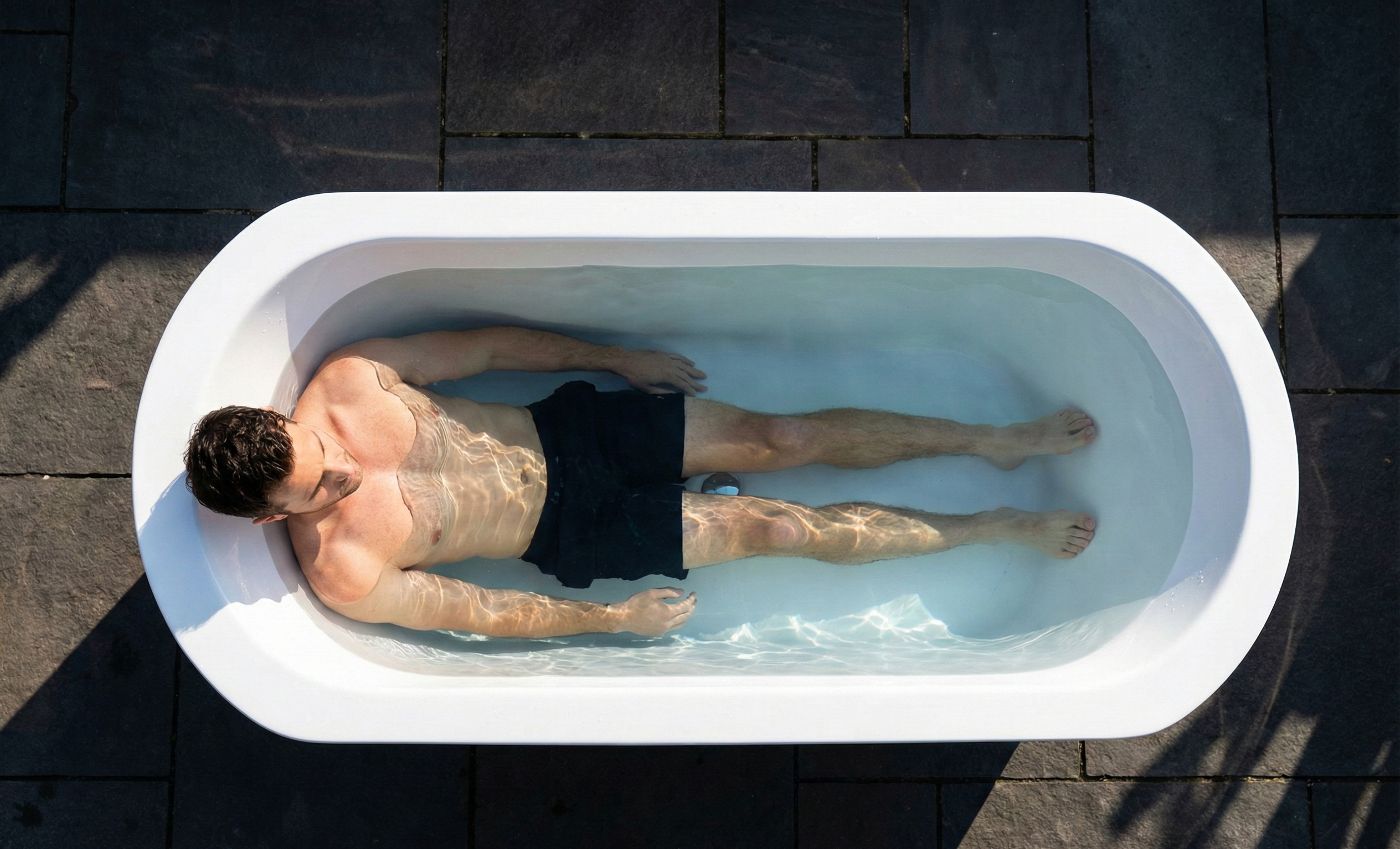 Person lying in a white bathtub outdoors on a dark tiled floor